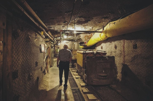 An elderly man walks through a dimly lit mine tunnel with mining carts on the tracks.