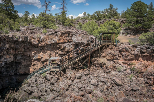 A wooden bridge spans a rocky landscape, leading to a deep crevice surrounded by trees and blue skies.
