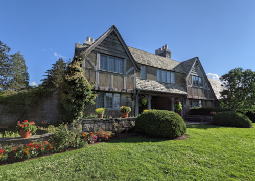 A charming two-story house with a stone facade, surrounded by lush greenery and colorful flower pots under a clear blue sky.