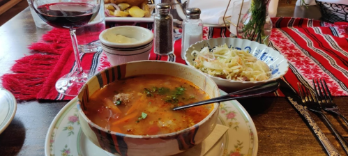 A bowl of soup with vegetables and meat, served with a side of coleslaw, wine, and a rustic table setting.