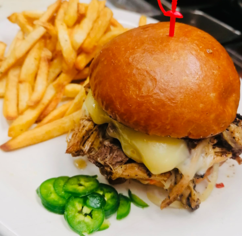 A pulled pork sandwich with melted cheese, served with crispy fries and sliced jalapeños on a white plate.