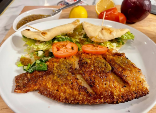 A plate featuring grilled fish with a spicy sauce, served with pita, lettuce, tomatoes, and lemon wedges.