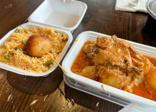 A takeout meal featuring fried chicken in sauce, rice with vegetables, and a cornbread muffin on a wooden table.