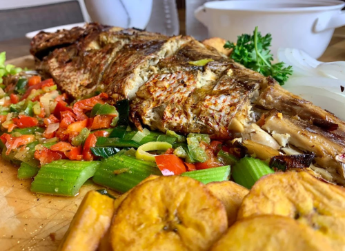 Grilled fish fillet served with sautéed vegetables, plantains, and fresh herbs on a wooden cutting board.