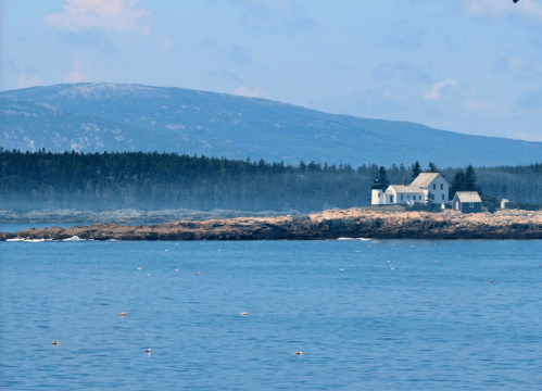 A serene coastal scene featuring a distant white house on a rocky shore, surrounded by blue water and misty mountains.