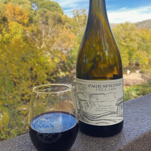 A bottle of Page Springs Cellars 2014 Petit Syrah next to a glass of red wine, with a scenic background of trees.