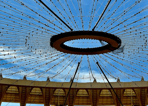 View of a circular, open-roof structure with radiating cables against a blue sky.