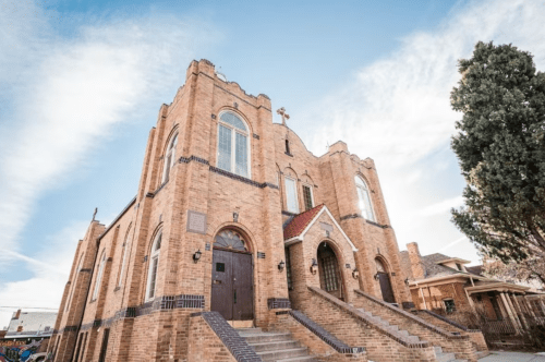 Historic brick church with a tall steeple, large windows, and steps leading to the entrance, under a blue sky.
