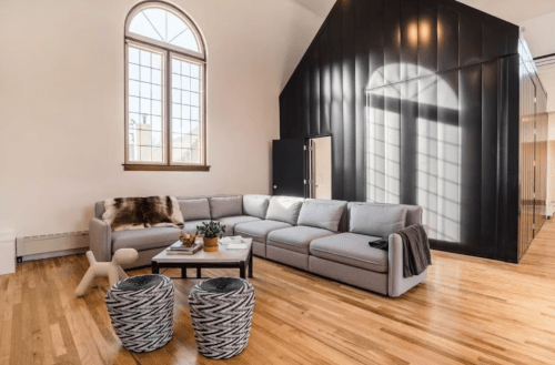 Modern living room with a large gray sectional, wooden floors, and stylish decor, featuring natural light from tall windows.