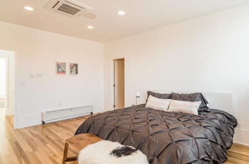 A modern bedroom featuring a large bed with a dark quilt, wooden floor, and minimalistic decor.