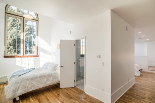 Bright, modern bedroom with a large window, wooden floors, and an open door leading to a bathroom.