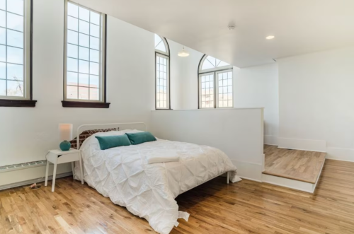 Bright, airy bedroom with a white bed, wooden floors, and large windows, featuring minimalist decor and natural light.