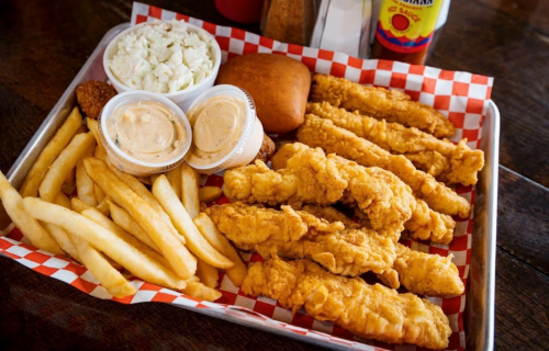 A tray of crispy fried chicken tenders, French fries, coleslaw, a roll, and dipping sauces on a checkered paper.
