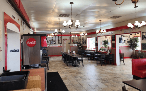 A cozy diner interior with red accents, a long table, booths, and vintage decor, featuring a Coca-Cola machine.