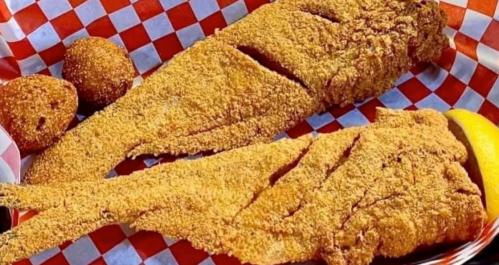 Two crispy fried fish fillets on a red and white checkered paper, accompanied by two golden hushpuppies and a lemon wedge.