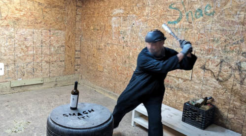 A person in a black outfit swings a bat at a wine bottle on a tire in a wooden shed.