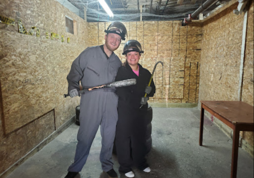 Two people in protective gear hold tools in a dimly lit, industrial-style room with wooden walls.