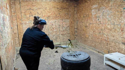 A person in a black outfit swings a bat at a tire labeled "SMASH IT" in a wooden room.