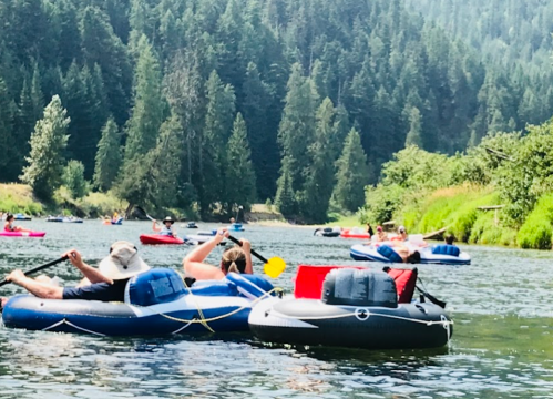 People in inflatable rafts paddle down a river surrounded by lush green trees and a sunny sky.