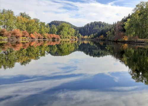 A serene lake reflects colorful autumn trees and a blue sky, surrounded by lush green hills.