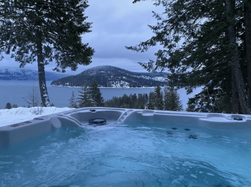 A hot tub overlooking a snowy landscape and lake, surrounded by evergreen trees under a cloudy sky.