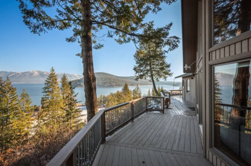 A scenic view from a wooden deck overlooking a lake surrounded by trees and mountains under a clear blue sky.