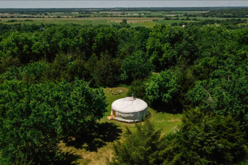 Aerial view of a yurt surrounded by lush green trees and open fields under a clear blue sky.