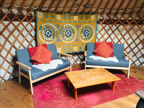 Cozy yurt interior with two blue sofas, orange cushions, a wooden coffee table, and a colorful wall tapestry.
