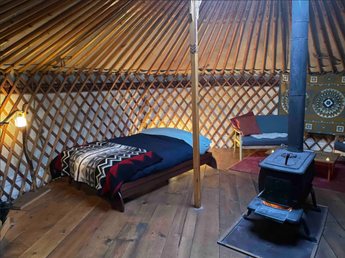 Cozy yurt interior with a bed, seating area, wooden floor, and a small stove, featuring a warm, rustic atmosphere.