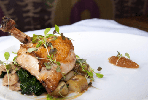 A plated dish featuring roasted chicken leg on a bed of greens and potatoes, garnished with microgreens and a sauce.