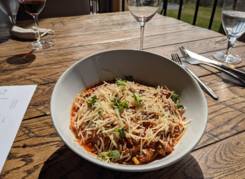A bowl of pasta topped with cheese and herbs, set on a wooden table with wine glasses in the background.