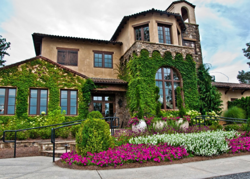 A charming stone and stucco house adorned with greenery and colorful flower beds in front.