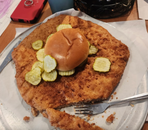 A large, crispy fried chicken sandwich topped with pickles on a metal plate, with a fork beside it.