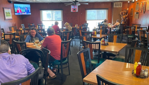 A spacious restaurant interior with wooden tables, chairs, and a TV, featuring diners enjoying their meals.