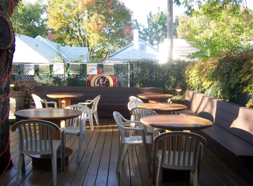 Outdoor seating area with wooden tables and chairs, surrounded by greenery and a fence, on a sunny day.