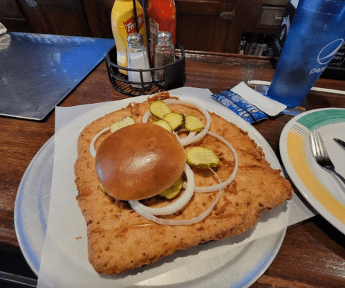 A large fried chicken sandwich with pickles and onions on a plate, served in a casual dining setting.