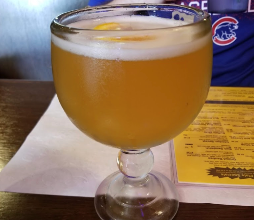 A glass of golden beer with a slice of lemon, sitting on a table with a menu in the background.