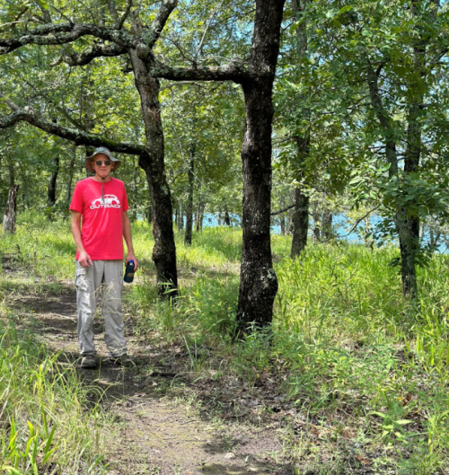 A person in a red shirt stands on a trail surrounded by trees and tall grass, with a body of water visible in the background.