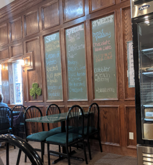 A cozy restaurant interior featuring wooden walls, a chalkboard menu, and empty tables with black chairs.
