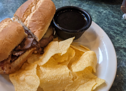 A plate with a sandwich filled with meat and a side of chips, accompanied by a small cup of dipping sauce.