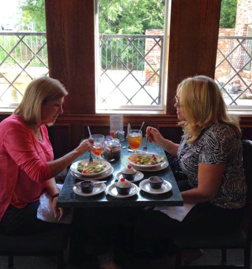 Two women sit at a table in a restaurant, enjoying meals and drinks, with a view of greenery outside.