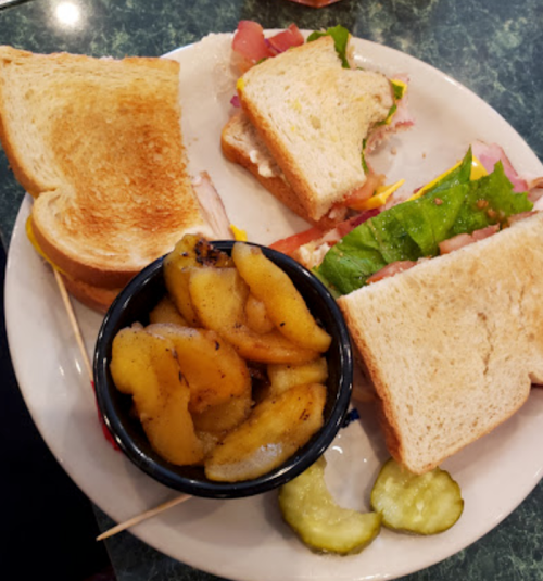 A plate with two toasted sandwiches, pickles, and a bowl of sliced apples.