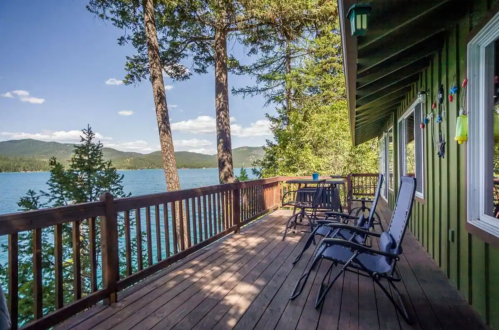 A wooden deck with chairs overlooks a serene lake surrounded by trees and mountains under a clear blue sky.