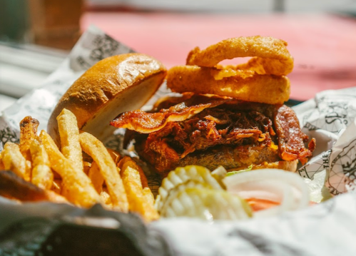 A close-up of a burger topped with bacon and onion rings, served with fries and pickles on a patterned paper.