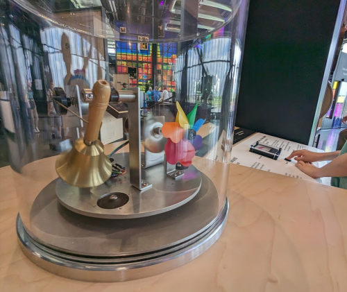 A glass display case containing a mechanical device with colorful components and a bell, on a wooden surface.