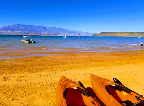 Two orange kayaks on a sandy beach with boats and mountains in the background under a clear blue sky.