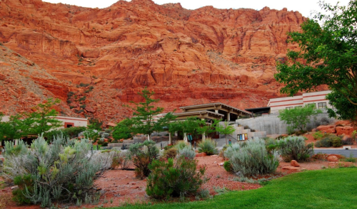 A desert landscape featuring red rock formations, greenery, and a modern building nestled at the base of the cliffs.