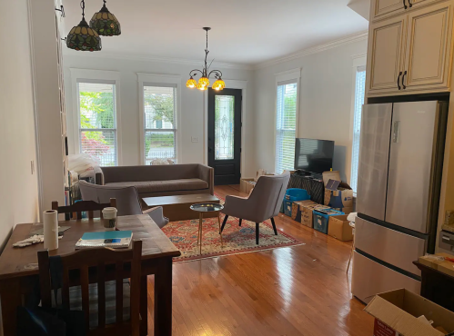 A cozy living room with a sofa, chairs, a dining table, and a TV, surrounded by boxes and natural light from windows.