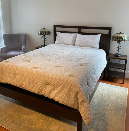 A cozy bedroom featuring a bed with a light quilt, two bedside tables, and a chair with stained glass lamps.