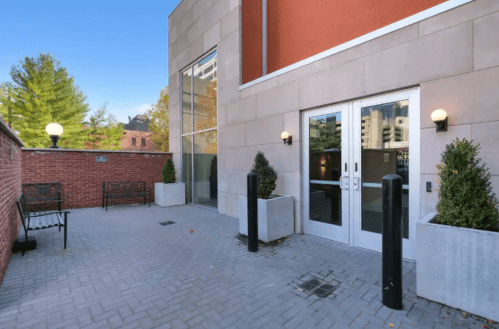Exterior view of a modern building entrance with glass doors, planters, and a bench in a landscaped area.